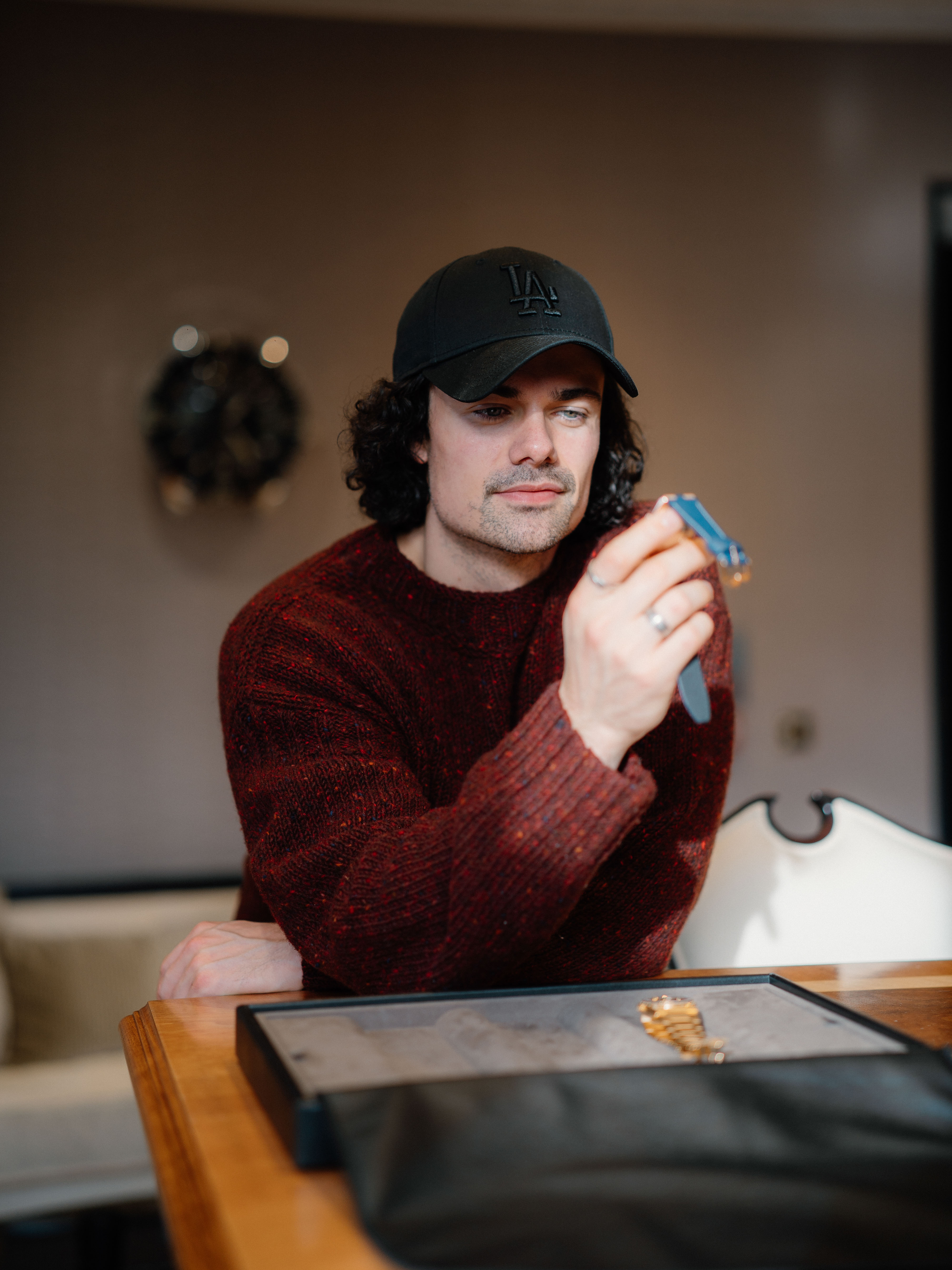 A man in a red sweater and black baseball cap looks at a watch with a blue strap that he is holding